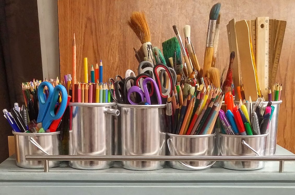 A collection of art supplies in buckets including scissors, colored pencils, rulers, paintbrushes, markers.