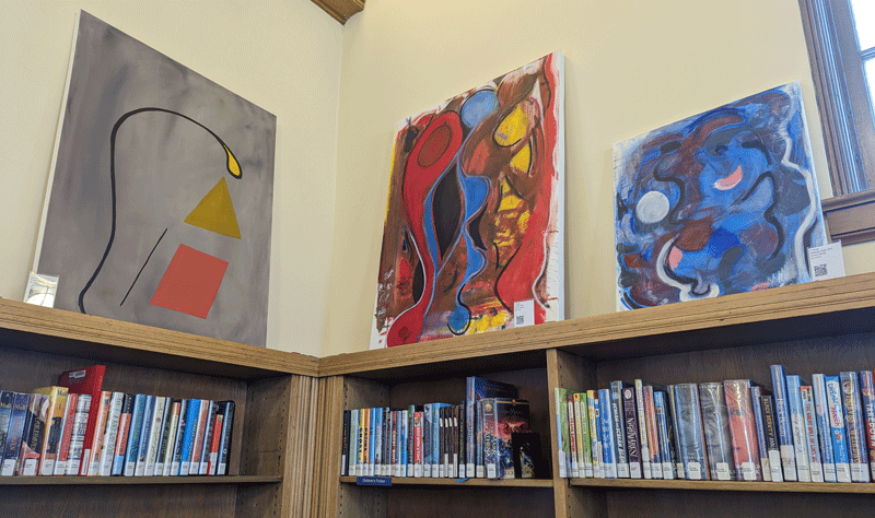 Three paintings by Chris Brown above a bookshelf at Maze Branch Library