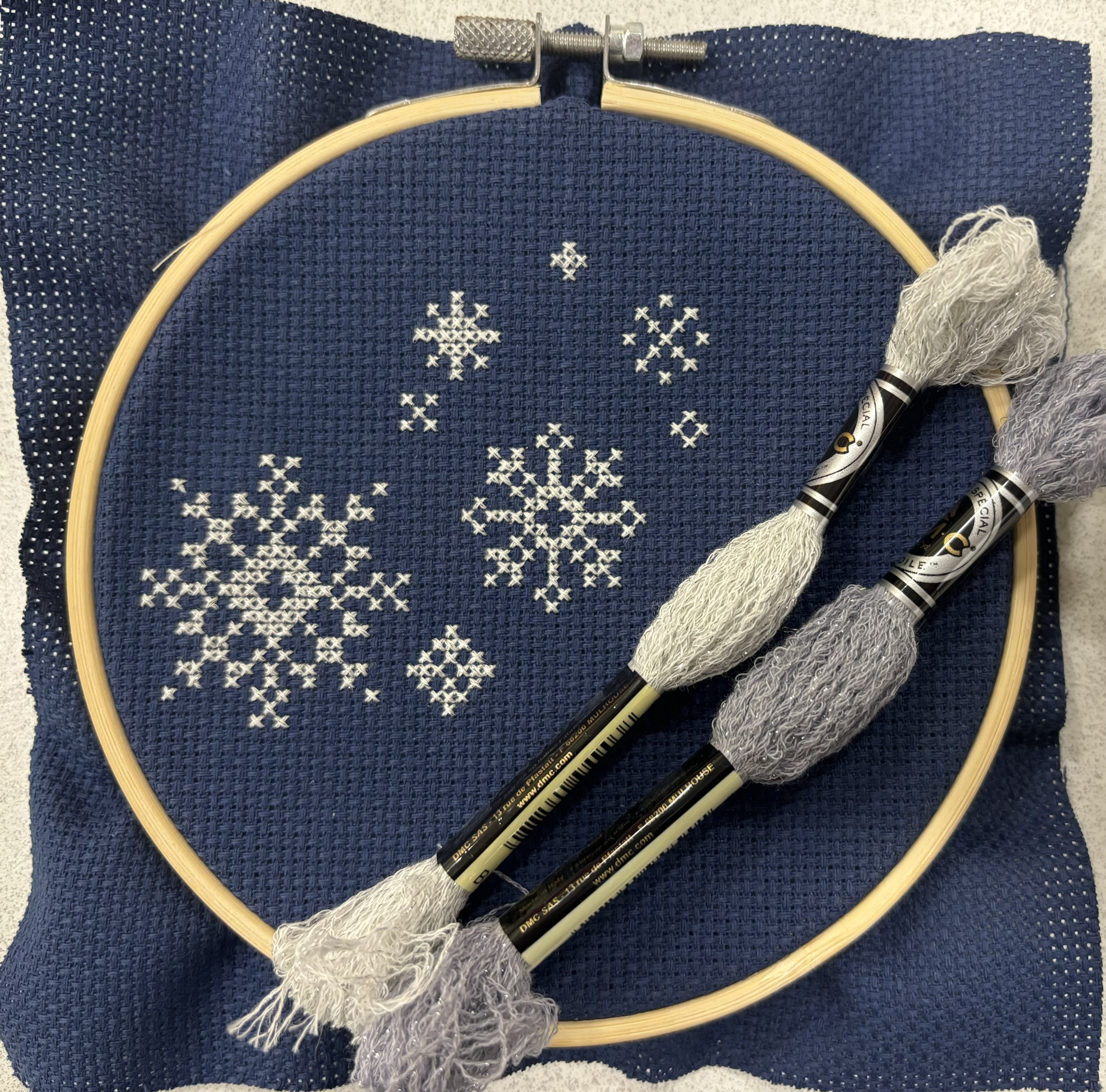 Dark blue 14 count aida in a wooden embroidery hoop. Seven white snowflakes are cross stitched on the fabric. One white skein and one gray skein of embroidery floss sit on top of the fabric and hoop.